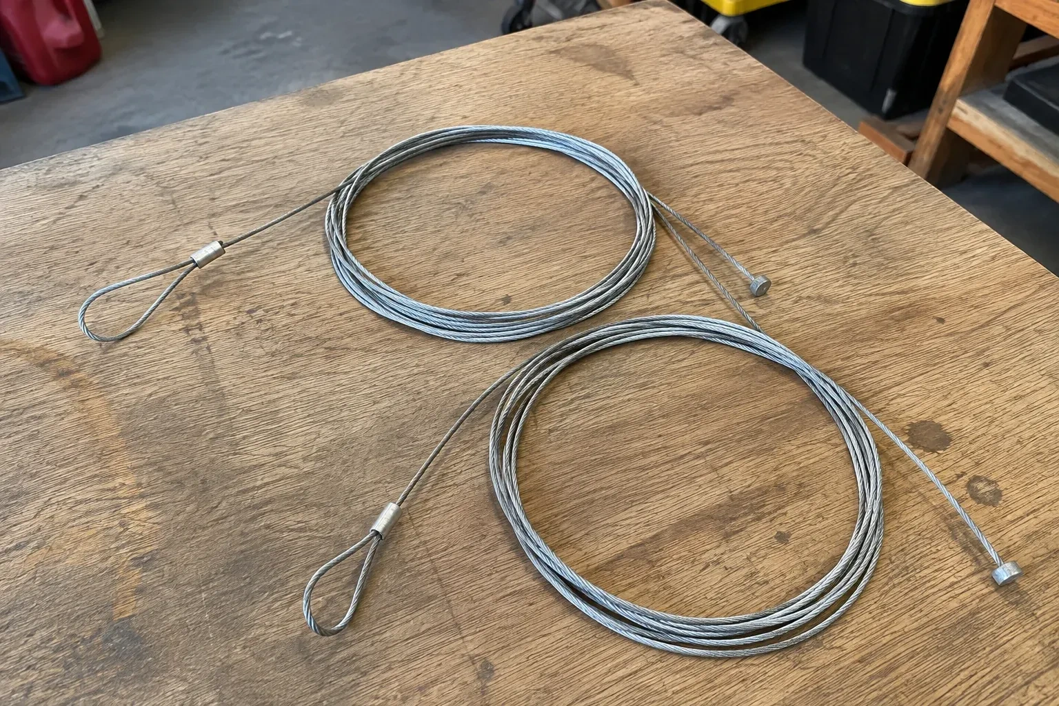 Two coiled steel cables with loop and stop ends on a workbench. — Garage Door Cable Replacement, Brant