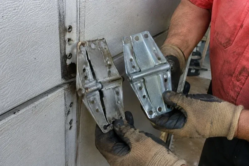 Gloved hands holding two metal garage-door hinges side by side, one discolored with a visible crack and the other bright silver. — Garage Door Hinge Replacement, Brant