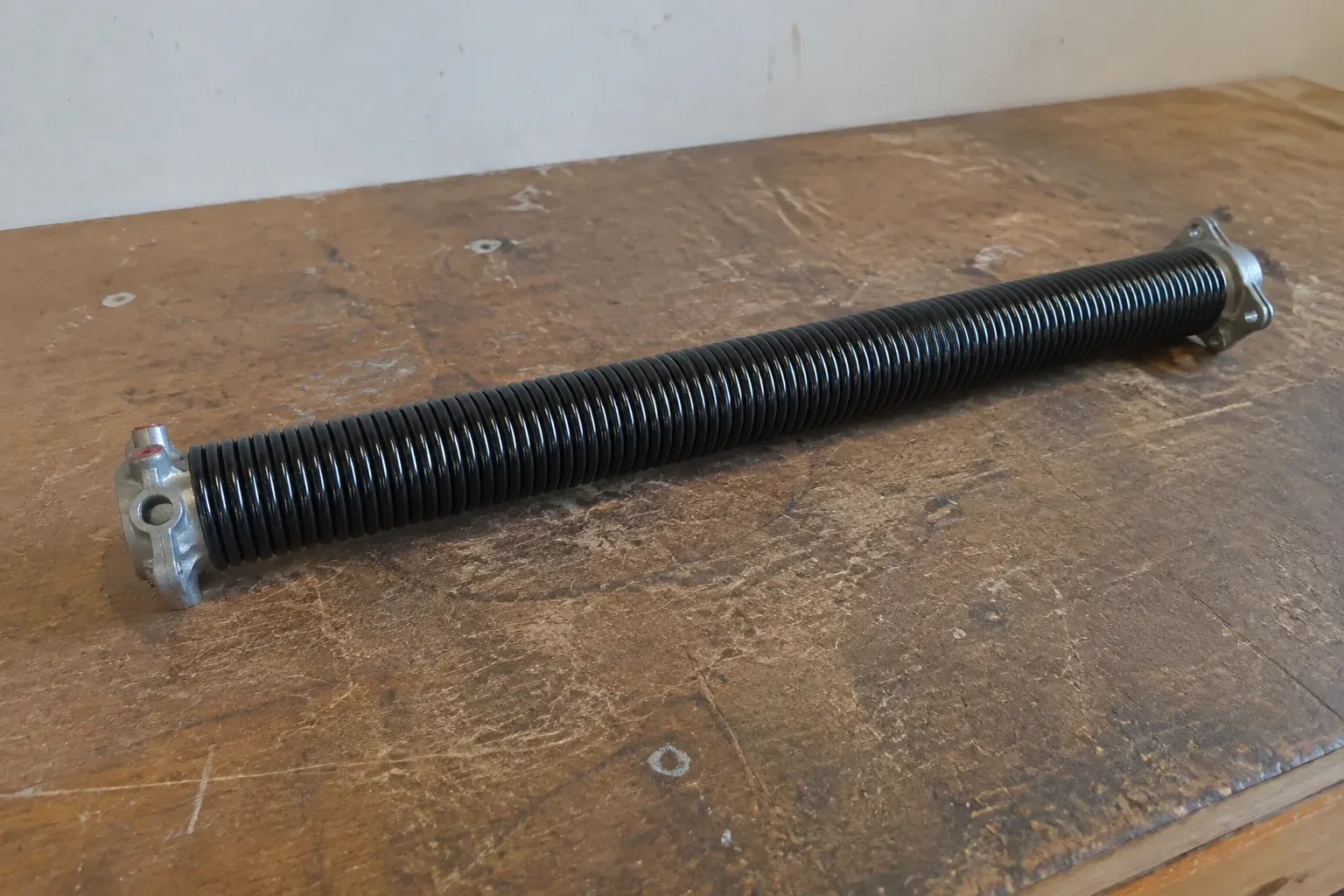 Detached black coil spring with metal end fittings on a workbench. — Torsion Spring Replacement, Brant